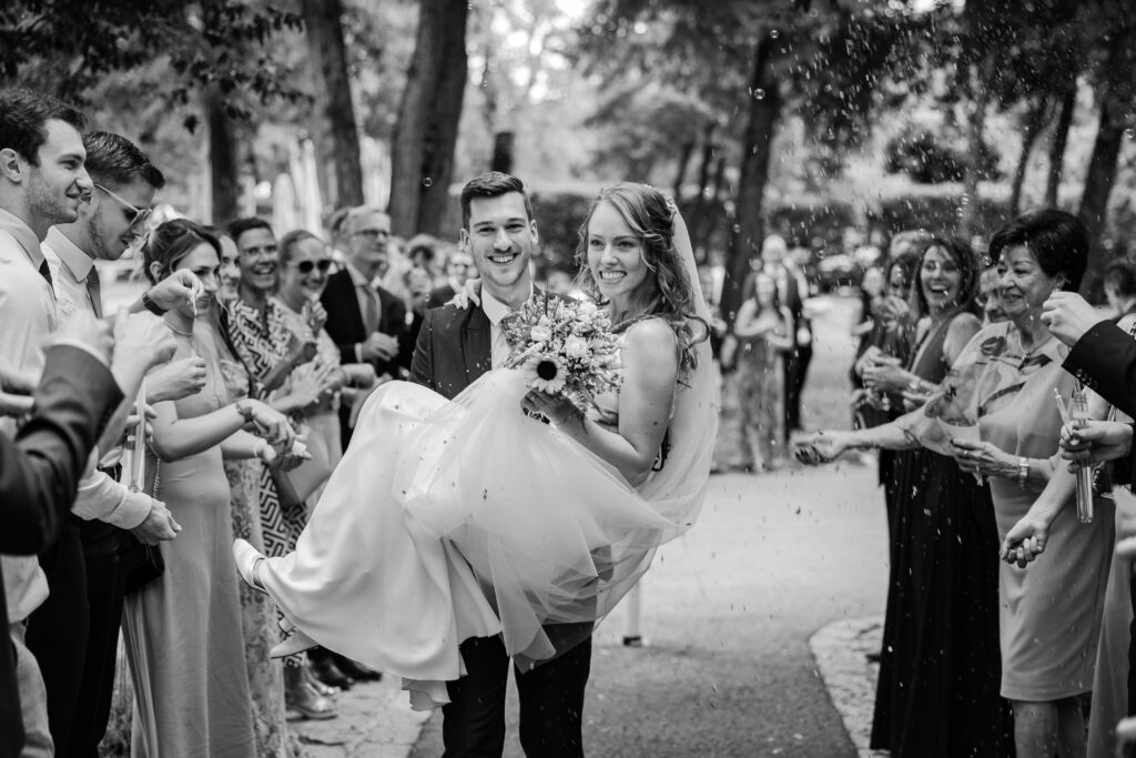 Bräutigam trägt Braut in den Armen durch den Spalier der Gäste