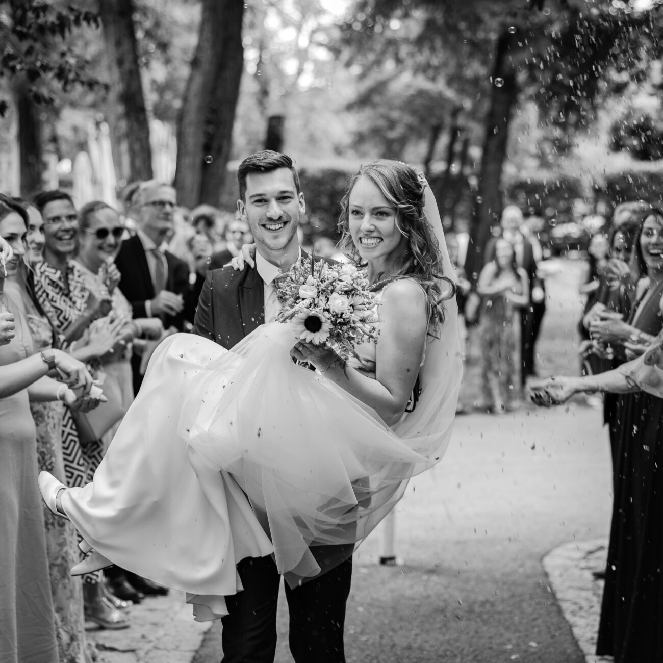 Bräutigam trägt Braut in den Armen durch den Spalier der Gäste